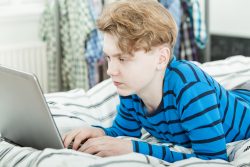 Trendy young teenage boy using a laptop computer as her relaxes on his bed at home doing his studies or browsing for recreation